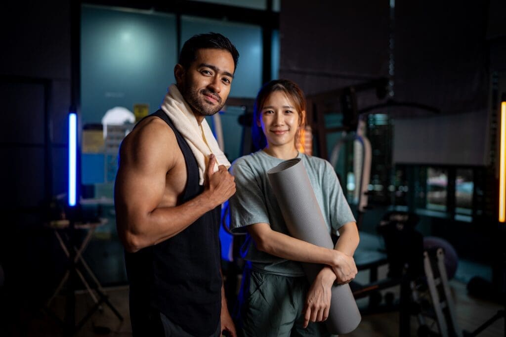 Two people in athletic clothing stand in a gym; one holds a towel over their shoulder, and the other holds a yoga mat.