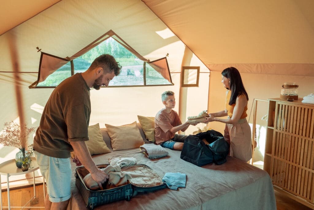 Three people are packing suitcases inside a tent with a bed, wooden shelves, and natural light from a window.